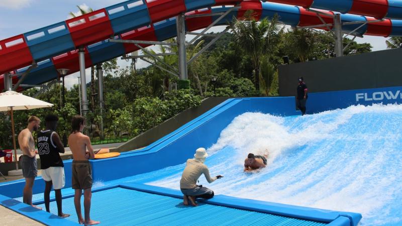 Гости аквапарка катаются на волновом аттракционе FlowRider: один человек стоит на доске на искусственной волне, а трое других ждут своей очереди на синем павильоне. На заднем плане видна конструкция водной горки и тропическая растительность.