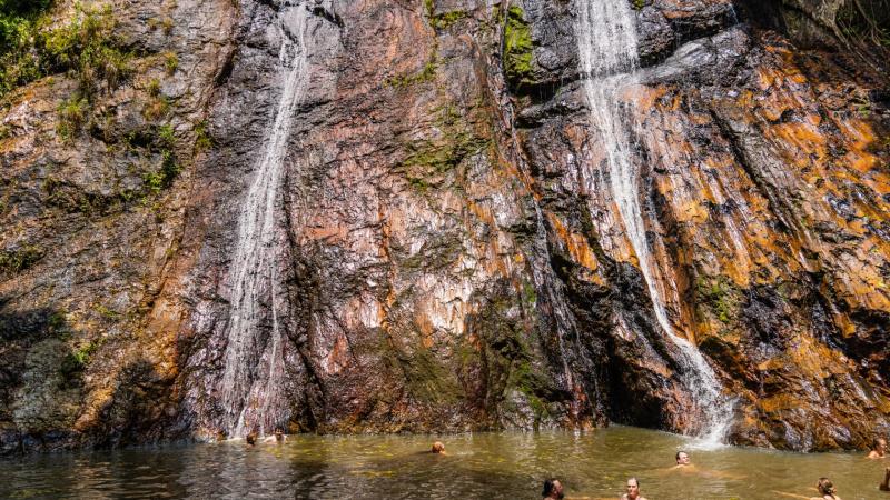 Два небольших водопада стекают по влажному скальному обрыву в природный бассейн с мутной водой, где купаются люди.