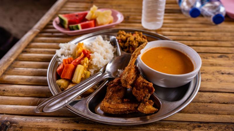 Металлический поднос с тайской едой: рис, жареное мясо, овощи и суп в белой миске. Рядом на бамбуковом столе — розовая тарелка с ломтиками арбуза и ананаса, пластиковая бутылка с водой и бутылки с синими крышками.