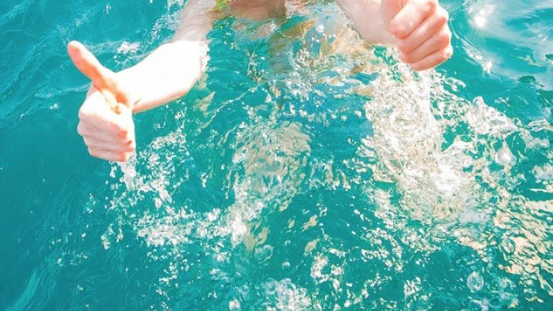 Руки человека, выходящего из бирюзовой воды, дают жест «большой палец вверх» и сжимают кулак. Вода вокруг рук мутная и пенистая, отражая солнечный свет.
