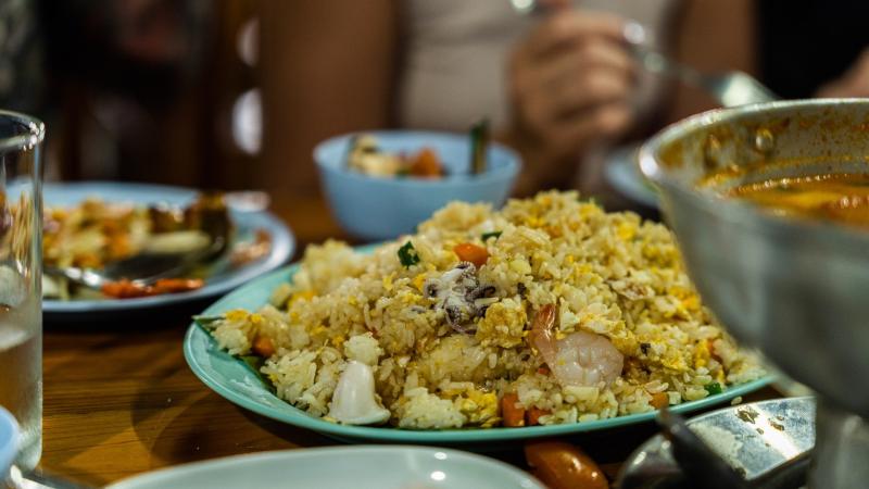 На деревянном столе в ресторане стоит тарелка с рисом, смешанным с яйцом, креветками и овощами. Рядом видны другие блюда, включая суп в металлической кастрюле, и бокал с напитком.