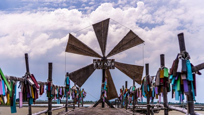 Деревянная ветряная мельница с надписью "WINDMILL" на мосту, окруженная деревянными столбами, на которых развешаны разноцветные ленточки. На заднем плане видны водная гладь и облачное небо.