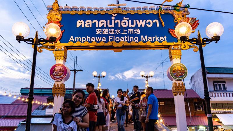 Группа туристов стоит под украшенным входным шлагбаумом с вывеской "Amphawa Floating Market" на тайском, китайском и английском языках. На заднем плане видны деревянные здания с красными крышами и фонари, освещающие вечерний рынок.