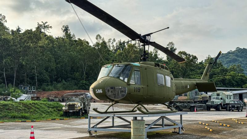 Зелёный военный вертолёт с номером 119 установлен на металлической подставке на открытой площадке. На заднем плане видны военные грузовики, лесной склон и другие объекты военной техники.
