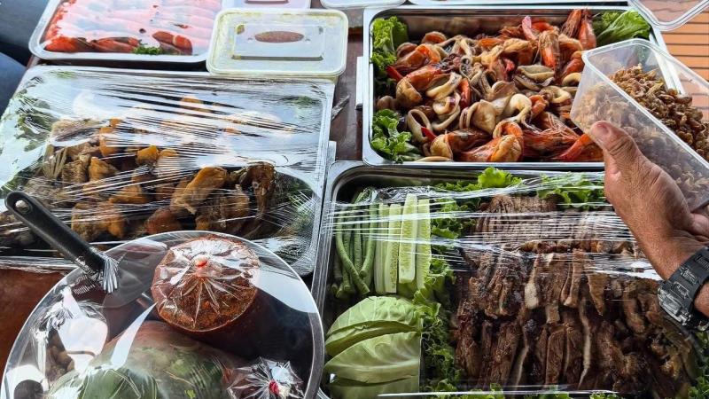 Разнообразные блюда тайской кухни на столе: жареные морепродукты с креветками и кальмарами, нарезанное мясо, свежие овощи, закуски и соусы в контейнерах. Рядом видна рука человека с часами, держащая прозрачную упаковку с едой.