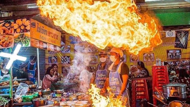 Уличная кухня в Паттайе: повара в масках и фартуках готовят блюда на открытом огне, над сковородой взлетает огромный языки пламени. На заднем плане видны посетители, меню с фотографиями еды и вывеска с тайской надписью.