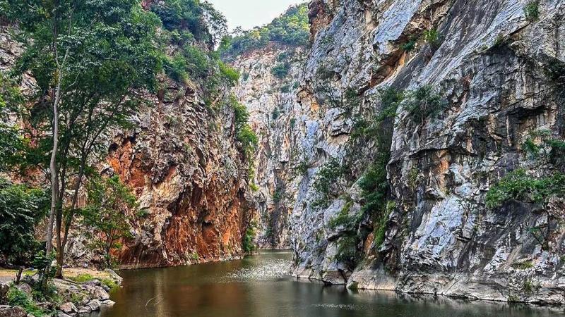 Узкая река с спокойной водой течет между высокими отвесными скалами, покрытыми трещинами и поросшими зеленью. На переднем плане видны деревья и каменистый берег, а в глубине ущелье сужается.