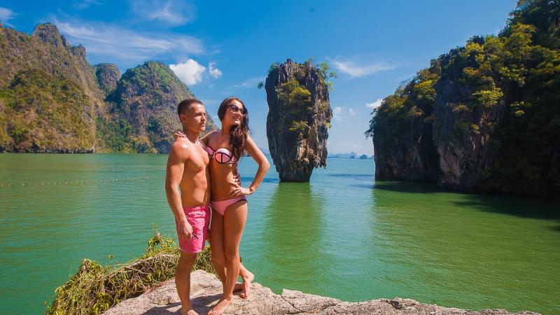 Пара фотографируется на камне у воды, за ними скала Ко Тапу и острова залива