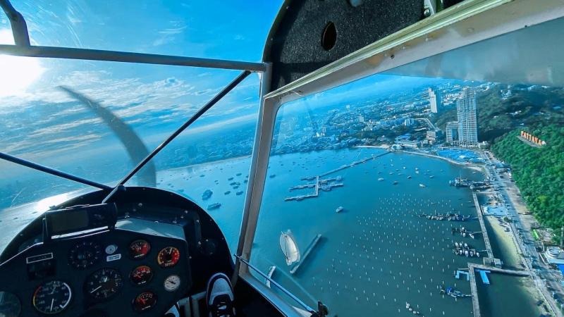 Вид из кабины самолёта на побережье Паттайи: голубое море с лодками, городские постройки вдоль берега и зелёные холмы на заднем плане. На переднем плане видна приборная панель и пропеллер.