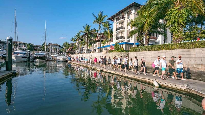 Гости идут по набережной Royal Phuket Marina к катамарану Sawanu вдоль яхт и пальм