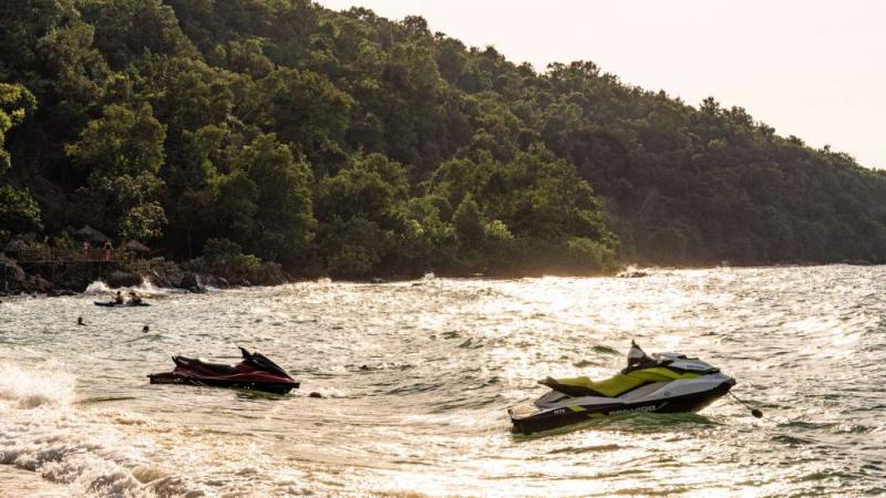 Два водных мотоцикла на воде у берега, покрытого густой растительностью. На заднем плане видны люди в воде и скалистый берег.