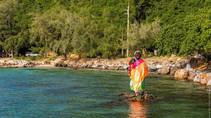 Женщина в ярком традиционном одеянии стоит на камнях в мелкой воде у берега. На заднем плане видны деревья, скалы и прибрежная полоса с автомобилями.