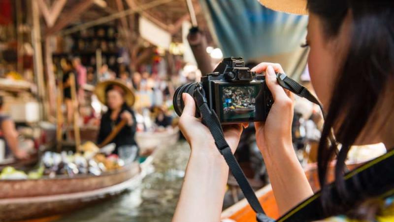 Туристка фотографирует на цифровой фотоаппарат сцены на плавучем рынке в Паттайе. На заднем плане видны лодки с товарами и местные продавцы в традиционных соломенных шляпах.