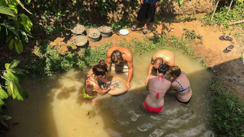Группа туристов в купальниках стоит по пояс в мутной воде природного бассейна, промывая золото в специальных поддонах. Рядом на берегу лежат корзины с песком и сланцами.