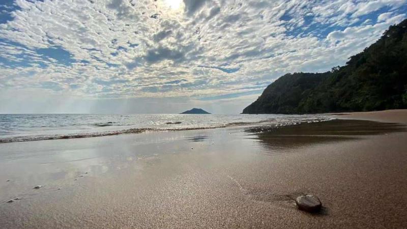 Песчаный пляж с мелкой галькой и спокойной водой, отражающей облачное небо. На заднем плане видны покрытые зеленью холмы и небольшой остров в море.