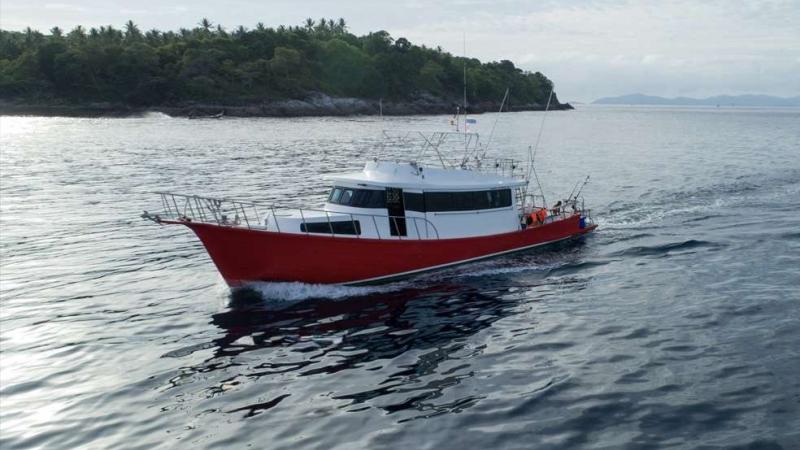Красно-белое рыболовное судно движется по спокойной воде, оставляя за собой небольшой след. На заднем плане виден покрытый густой растительностью берег острова в районе Пхукета.