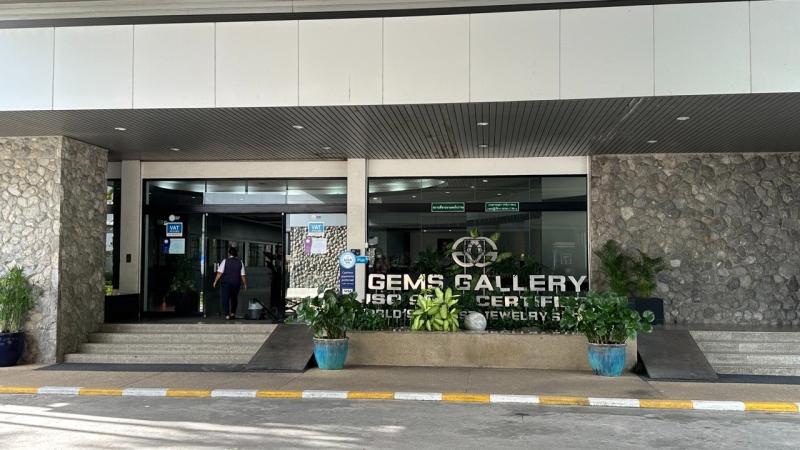 Вход в здание ювелирной фабрики Gems Gallery на Пхукете: стеклянные двери, каменные стены и ступени. Перед входом стоят вазоны с растениями, на фасаде видна вывеска с логотипом и надписью "GEMS GALLERY".