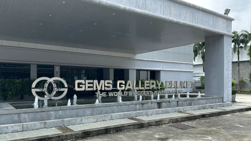 Вход в ювелирный магазин Gems Gallery на Пхукете с вывеской "The World's Biggest Jewelry Store" и логотипом. Перед зданием расположена декоративная фонтанная чаша с несколькими струями воды, а справа видны пальмы.