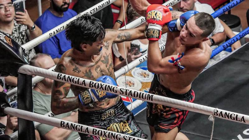 Два бойца в клинче у канатов с надписью Bangla Boxing Stadium, за рингом — зрители
