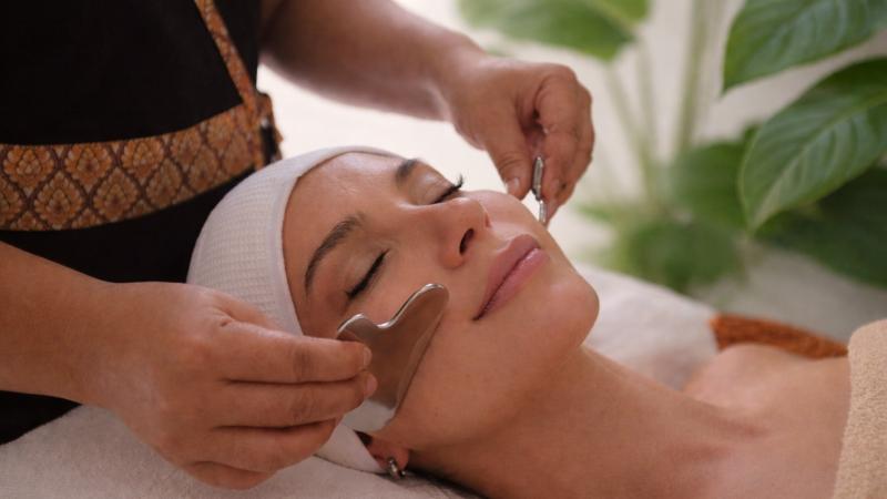 Уход Gua Sha: мастер ведёт металлическим скребком по лицу гостьи в белой повязке