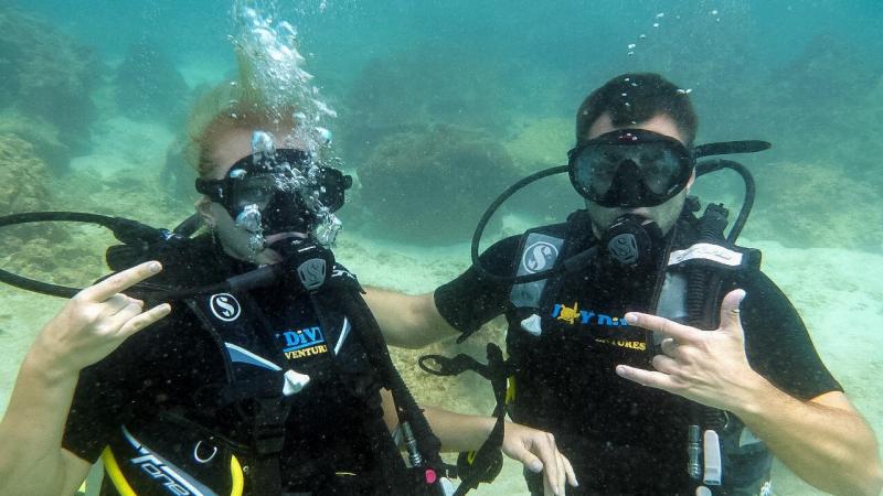 Два человека в снаряжении для дайвинга под водой на фоне песчаного дна и кораллов. Оба делают жест рукой, на груди видны логотипы дайвинг-клуба.