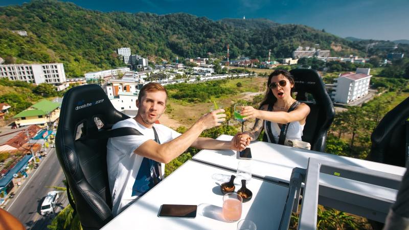 Гости чокаются коктейлями за столом Dinner in the Sky, внизу холмы и дома Карона