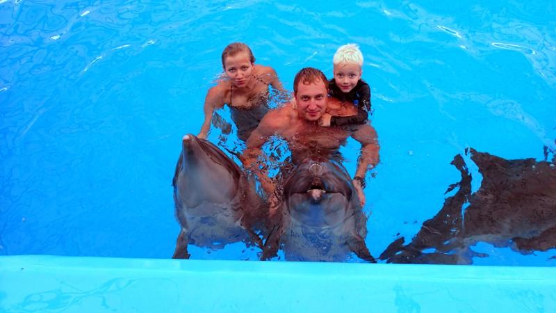 Мужчина с двумя детьми в воде бассейна дельфинария в Пхукете. Рядом с ними плывут два дельфина, один из которых находится прямо перед мужчиной.