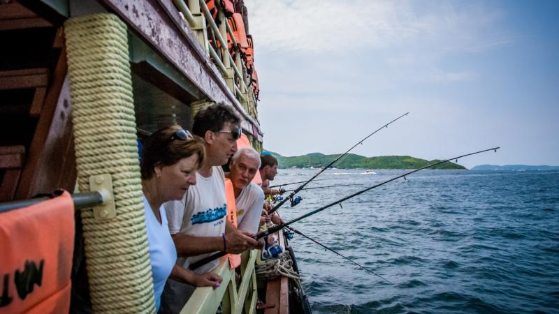Группа людей стоит на палубе лодки и держит удочки, нацеленные в море. На заднем плане виден зеленый остров, покрытый растительностью, под ясным небом.