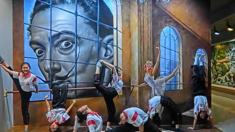 Девушки в спортивной форме позируют в различных позах перед 3D-мural с изображением лица, выглядывающего из-за решетки. На заднем плане видны архитектурные элементы и окна, создающие эффект глубины.