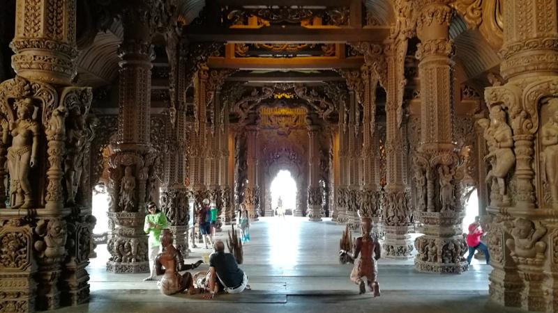 Внутренний вид зала Храма Истины в Паттайе с рядами intricately carved деревянных колонн, украшенных скульптурами, и светящимся проходом в глубине. На полу сидят несколько посетителей, наблюдающих за деталями интерьера.