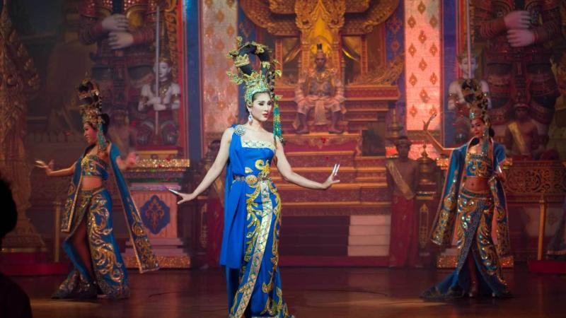 Три танцовщицы в ярких синих и золотых костюмах с высокими головными уборами исполняют сцену на сцене. Фон украшен богатыми золотыми деталями и изображениями в тайском стиле, характерными для шоу в Паттайе.