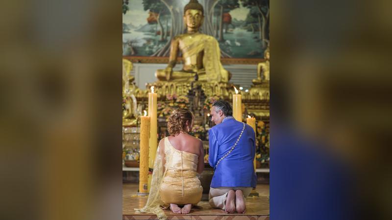 Пара в тайских нарядах на коленях перед статуей Будды и свечами — церемония в храме