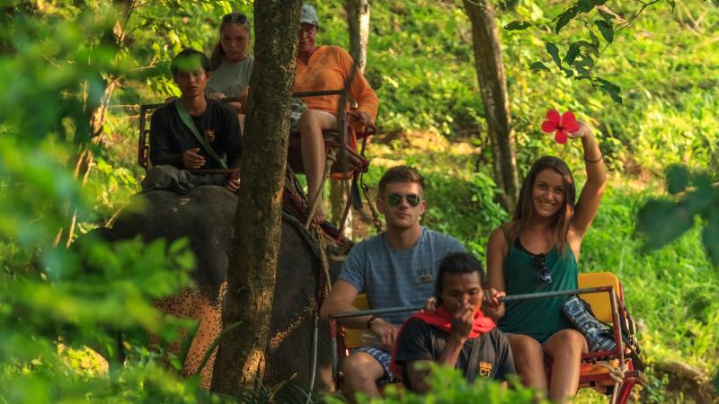 Пара в кресле на спине слона машет в камеру, впереди сидит погонщик — Kok Chang Safari