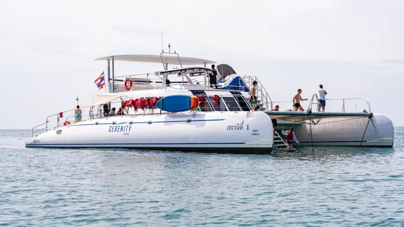 Белый катер-катамаран "Serenity" с пассажирами на борту и палубе, стоящий на воде. На корме виден флаг Таиланда, а на борту — спасательные круги и надувная лодка.