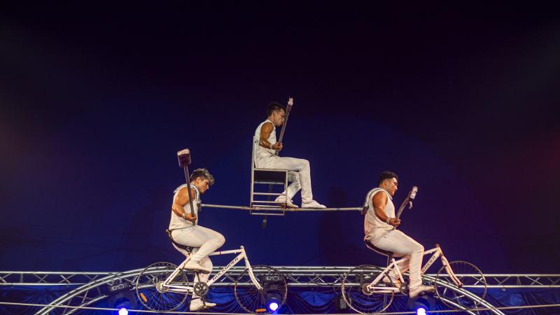 Два артиста на велосипедах едут по канату, на шесте между ними сидит третий на стуле