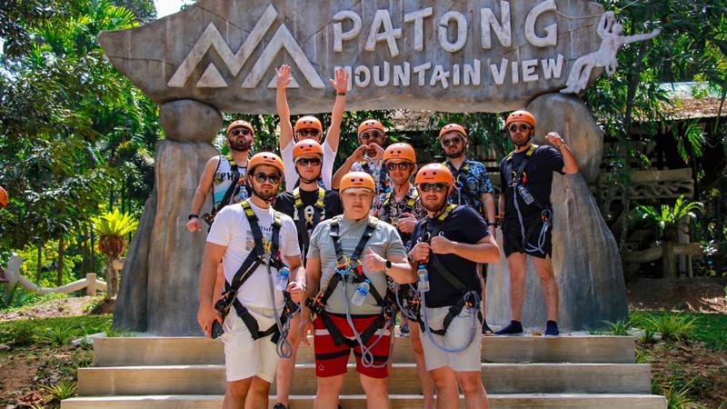 Гости в шлемах и страховочных системах у входа с вывеской Patong Mountain View перед трассой зиплайна