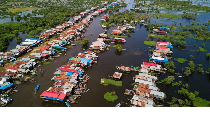 Вид сверху на плотное поселение на воде: ряды домов на сваях с красными и синими крышами тянутся вдоль узкой реки, окруженной густой зеленью. По воде расходятся круги от проплывающих лодок.