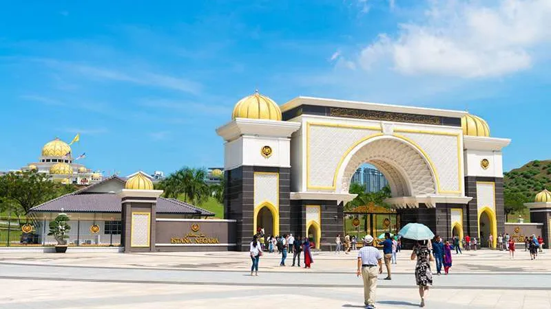 Большой арочный вход с золотыми куполами и надписью "ISTANA NEGARA" на стене. На переднем плане туристы гуляют по открытой площади, вдалеке видны здания с золотыми куполами и холмы на фоне голубого неба.