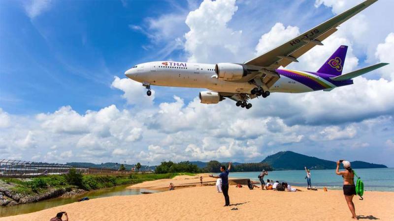 Самолёт авиакомпании Thai Airways с опущенными шасси летит низко над пляжем. На песчаном берегу люди фотографируют пролетающий лайнер на фоне моря и холмов.