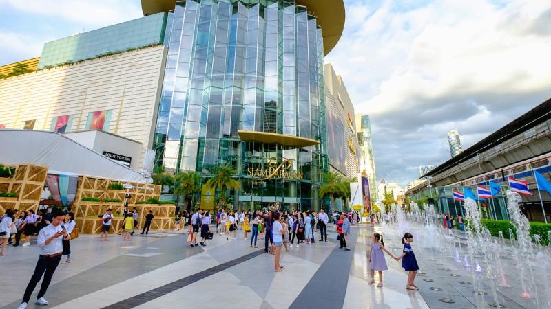 Пешеходная площадь перед стеклянным зданием с вывеской "Siam Paragon" в Бангкоке. На переднем плане дети играют у фонтанов, в центре толпа туристов, справа видны тайские флаги и крытая галерея.