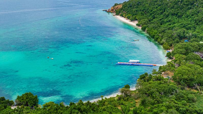 Вид с высоты на изумрудно-бирюзовое море у берега, покрытого густой зелёной растительностью. У берега виден плавучий причес с навесом, а в воде — несколько небольших лодок.