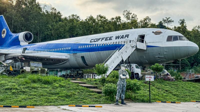 Бывший пассажирский самолет с надписью "COFFEE WAR" установлен на травяном склоне среди деревьев и используется как кафе. Рядом с ним стоят военные джипы, а на переднем плане — фигура солдата в камуфляже.