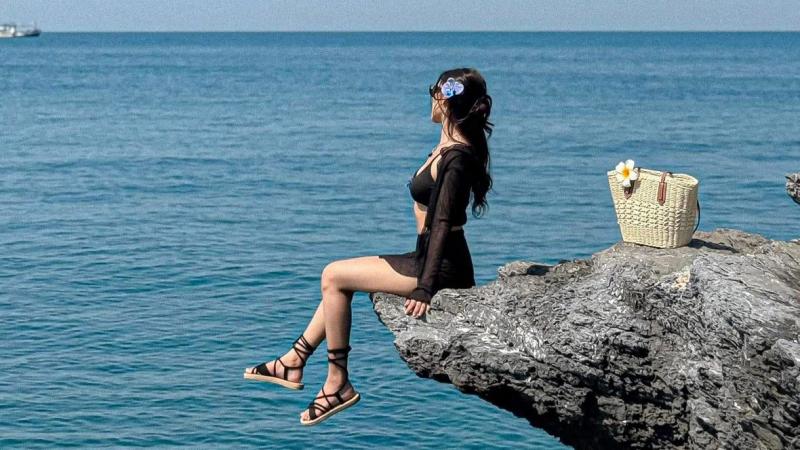 Девушка в черном купальнике и легком топе сидит на выступающей над водой скале, глядя на море. Рядом с ней на камне стоит плетеная корзина с белым цветком.
