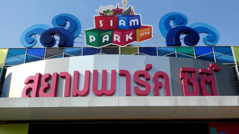 Фасад здания с большим красным таиландским текстом и надписью "SIAM PARK CITY" на разноцветных кубах. Над входом расположены декоративные синие волны и изображения тайских архитектурных элементов.