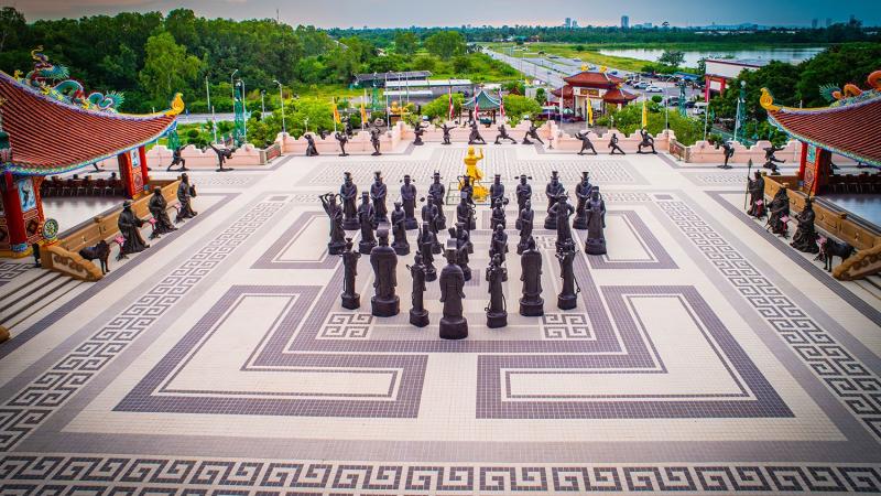 Большая открытая площадка с гигантской шахматной доской, на которой стоят фигуры в виде статуй. По периметру расположены дополнительные скульптуры, а на заднем плане видны традиционные азиатские здания с красными крышами и зеленые насаждения.