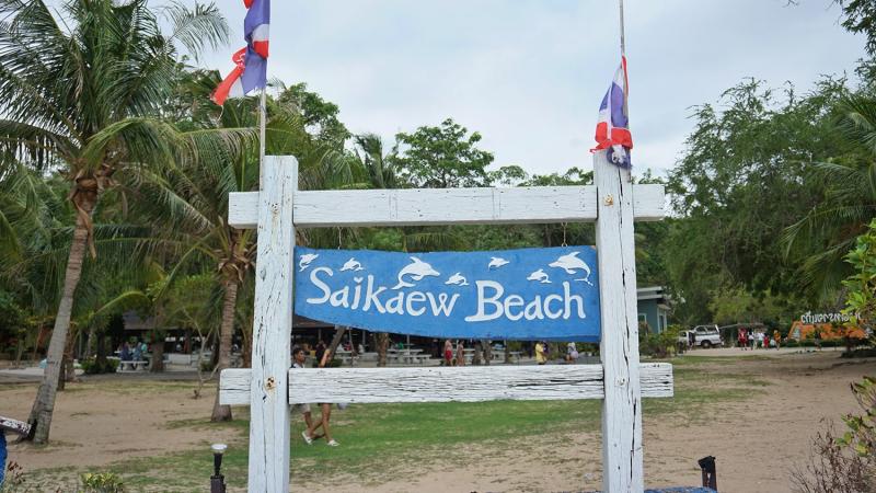 Белый деревянный знак с синей табличкой, на которой написано "Saikaeo Beach" и изображены дельфины. На столбах знака установлены флаги Таиланда. На заднем плане видны пальмы, деревья и песчаная территория пляжа.