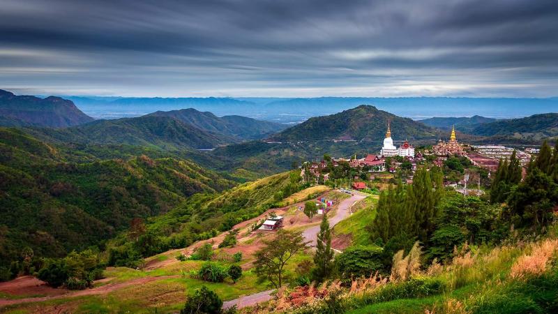 Панорама холмистого пейзажа с храмовым комплексом на вершине горы, включающим белую ступу и золотые шпильки. На переднем плане видны зелёные склоны с деревьями и дорожки, ведущие к храму, а на заднем плане — горные хребты под облачным небом.