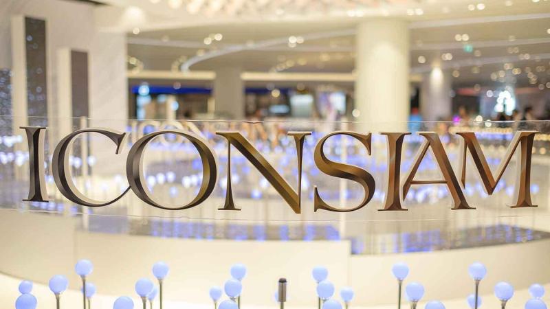 Золотые буквы надписи ICONSIAM на стеклянной поверхности на фоне размытого интерьера с колоннами и светящимися точками. Внизу видны декоративные голубые шары на тонких ножках.