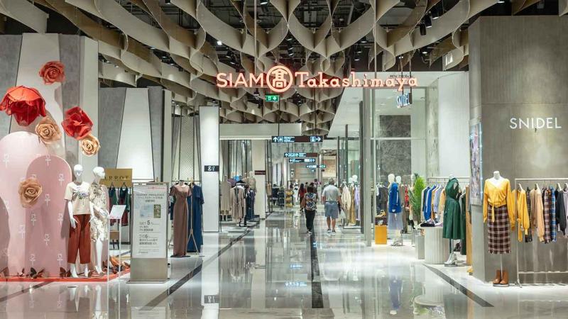 Внутренний вид современного торгового центра с вывеской SIAM Takashimaya, где расположены магазины одежды, включая SNIDEL. Просторный зал с глянцевым полом, манекенами в витринах и декоративными цветами.